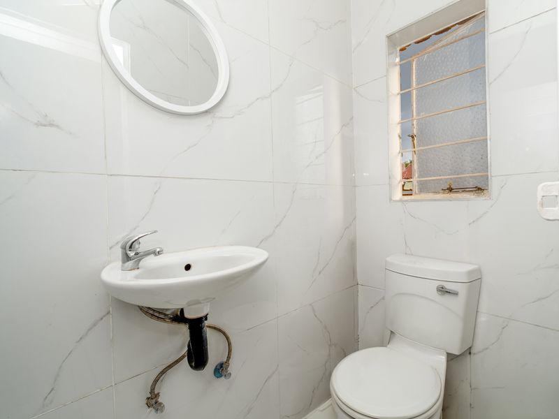 A compact bathroom featuring white marble-look wall tiles, a wall-mounted basin, and a standard toilet, requiring minor repairs to plumbing and window fixtures.