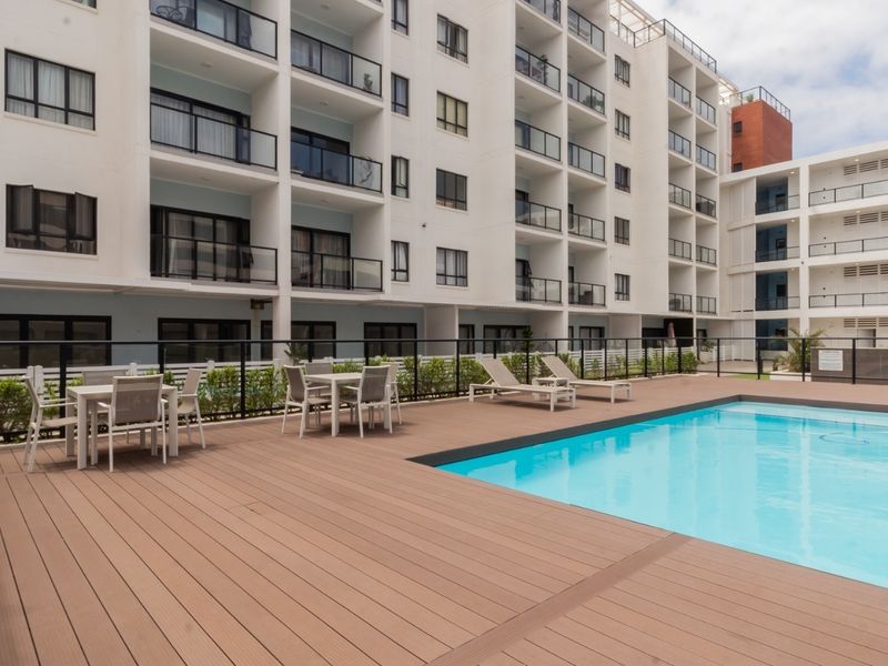 A well-maintained communal swimming pool area featuring modern composite decking, glass safety balustrades, and direct access to the apartment building complex.