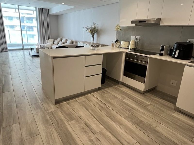 Modern open-plan kitchen and living area featuring sleek white cabinetry, stone countertops, integrated appliances, and wood-look tile flooring leading to a balcony.