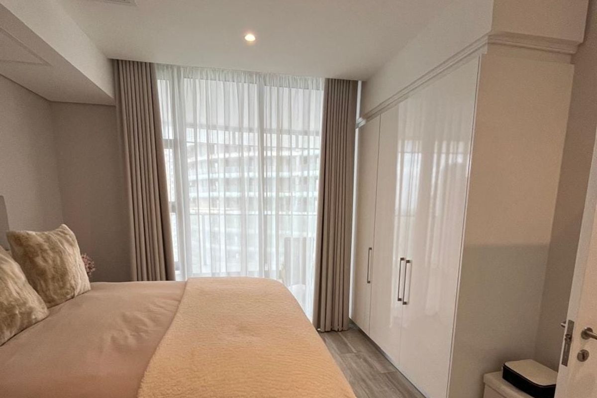 A well-maintained bedroom featuring modern built-in high-gloss wardrobes, recessed ceiling lighting, and neutral laminate flooring with floor-to-ceiling window treatments.