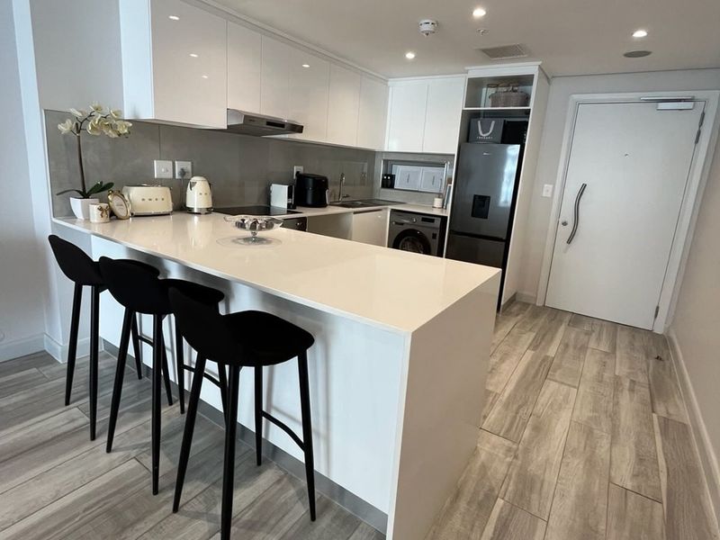 Modern kitchen featuring white gloss cabinetry, stone countertops, a breakfast bar, and wood-look tile flooring in an open-plan layout.