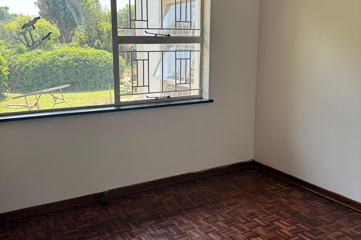 A bright, empty bedroom featuring well-maintained parquet flooring, a large window with security bars, and a neutral wall finish.