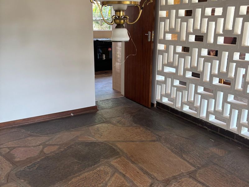 An empty living area featuring distinctive flagstone flooring, a decorative breeze block partition wall, and a timber ceiling with a vintage pendant light.