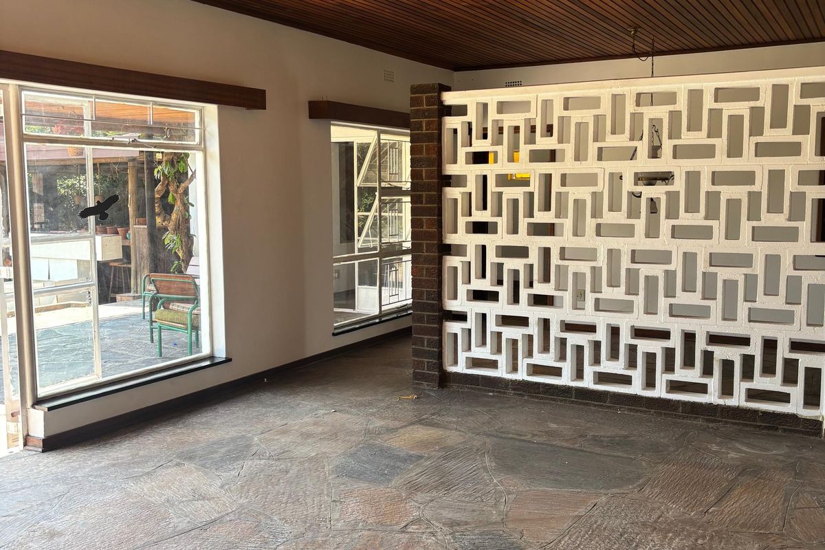Spacious living area featuring a unique timber slat ceiling, decorative breeze block partition wall, and rustic flagstone flooring with large windows.