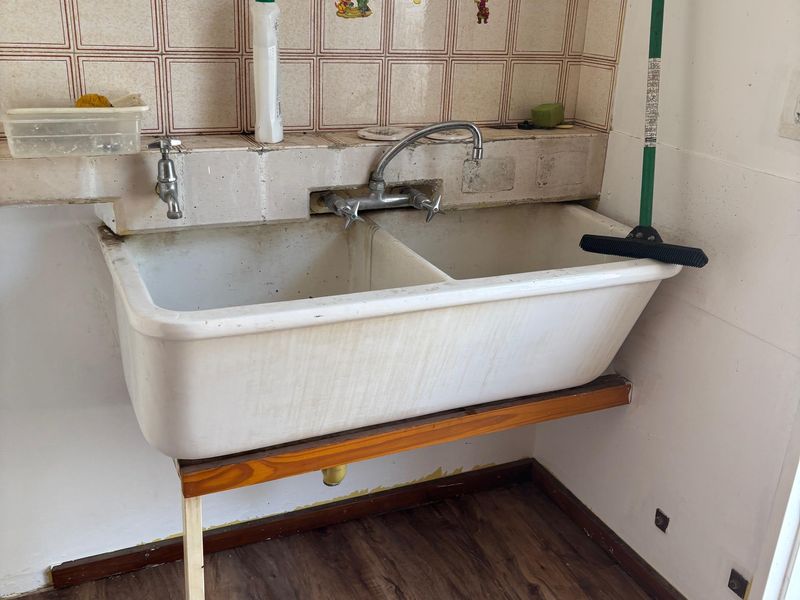 A functional laundry area featuring a double basin ceramic sink with wall-mounted taps and tiled splashback, set against a neutral wall finish.