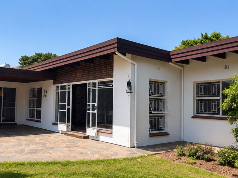 A well-maintained rear exterior view featuring a stone-paved patio, white rendered walls, brick accents, and secure metal-framed windows under a deep roof overhang.