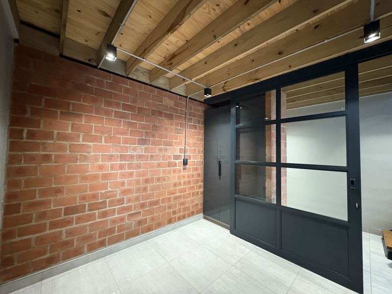 Modern industrial-style living space featuring exposed brick walls, timber ceiling beams, and a sleek black-framed glass partition door.