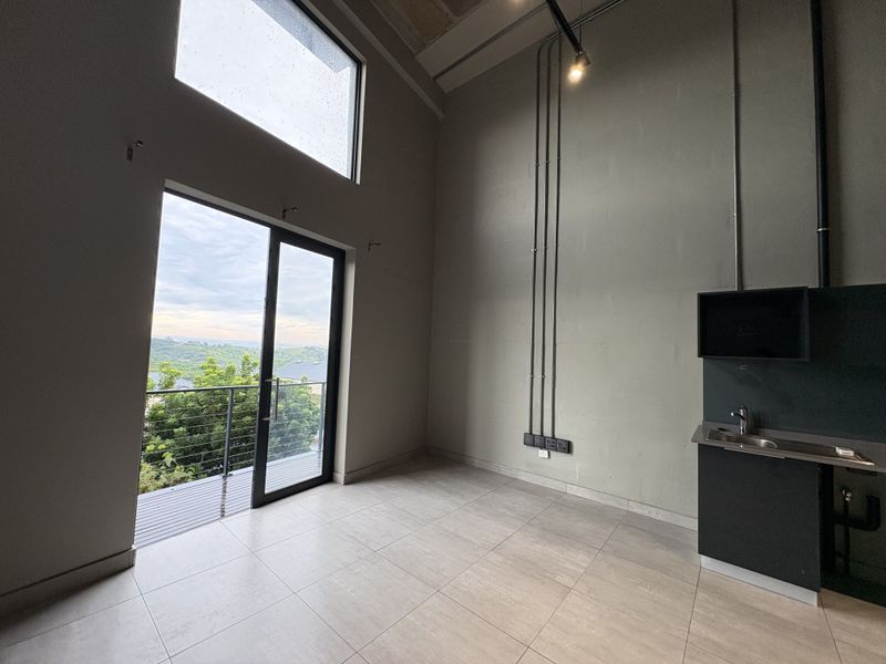 Modern open-plan studio space featuring high ceilings, exposed industrial conduit, tiled floors, a kitchenette, and a sliding door leading to a private balcony.