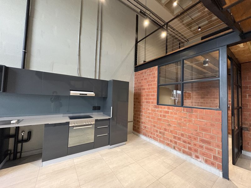 Modern industrial-style kitchen featuring sleek dark cabinetry, integrated appliances, exposed brickwork, and a mezzanine level with steel detailing.