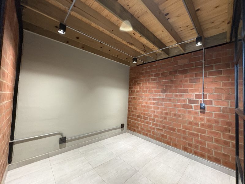 An empty industrial-style room featuring exposed brick walls, timber ceiling beams, track lighting, and modern tiled flooring.