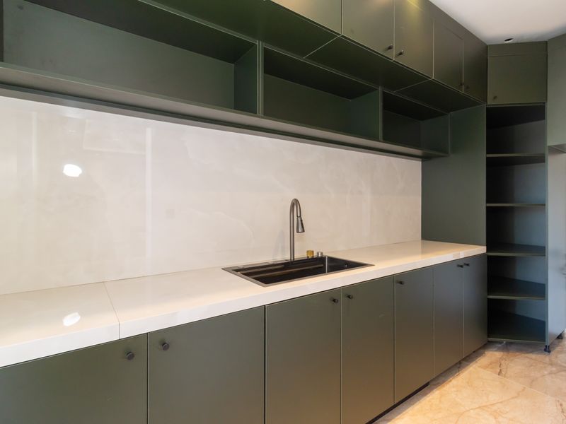 A modern scullery featuring dark green cabinetry, white stone countertops, an undermount sink with a gooseneck faucet, and integrated open shelving.