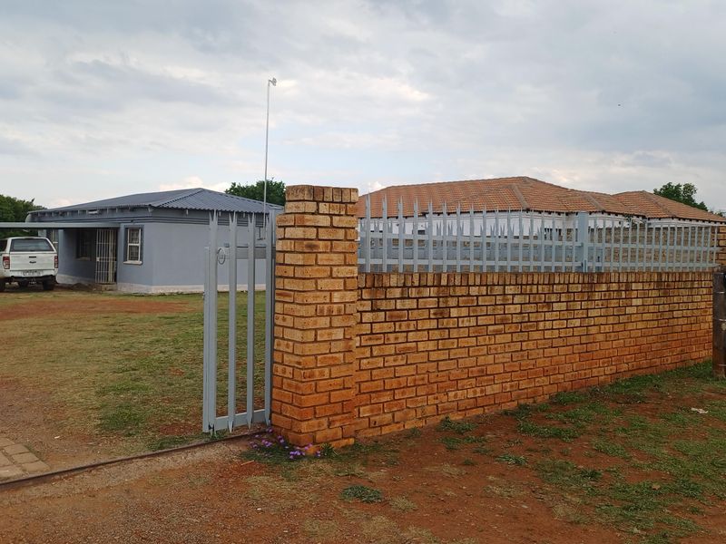 Front view of the property featuring a face brick boundary wall, steel security fencing, and a carport on a large open plot.