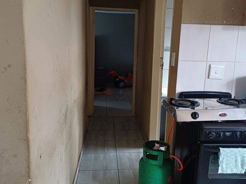 A kitchen area featuring tiled flooring, a gas stove, and tiled splashbacks, leading into a hallway. The walls show signs of wear and staining.