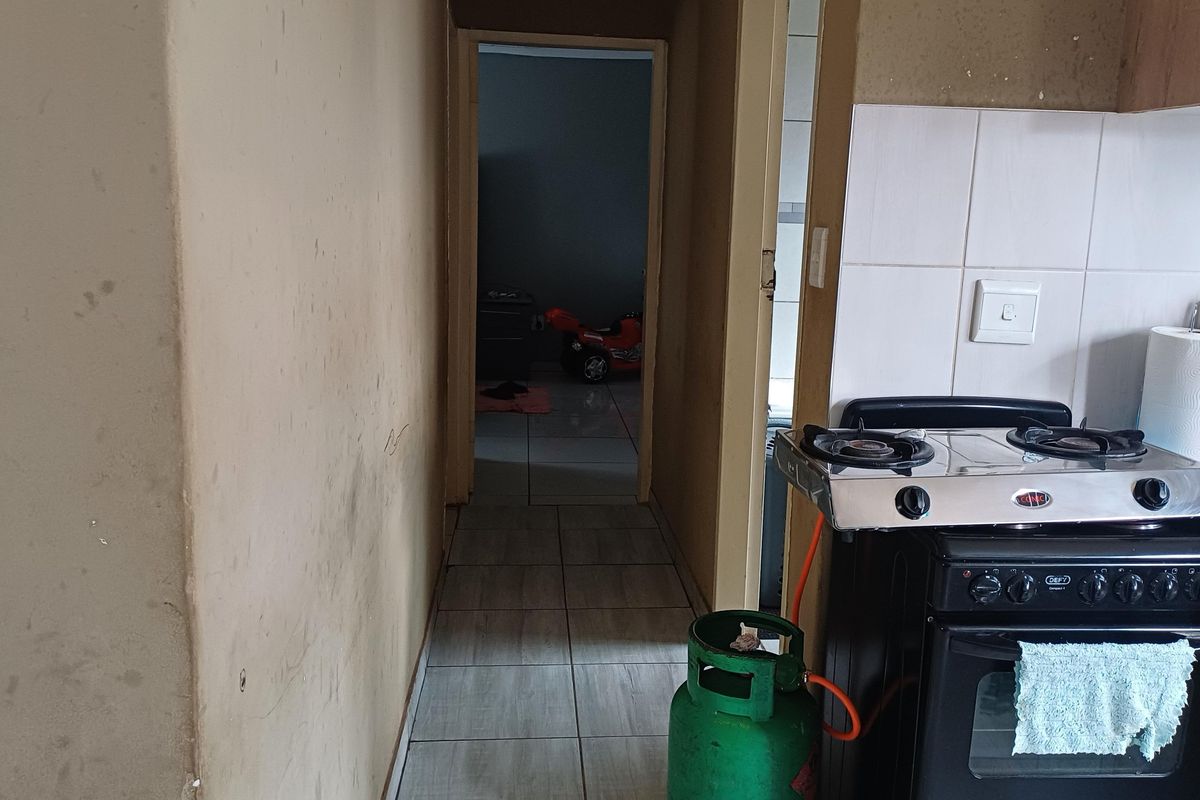 A kitchen area featuring tiled flooring, a gas stove, and tiled splashbacks, leading into a hallway. The walls show signs of wear and staining.