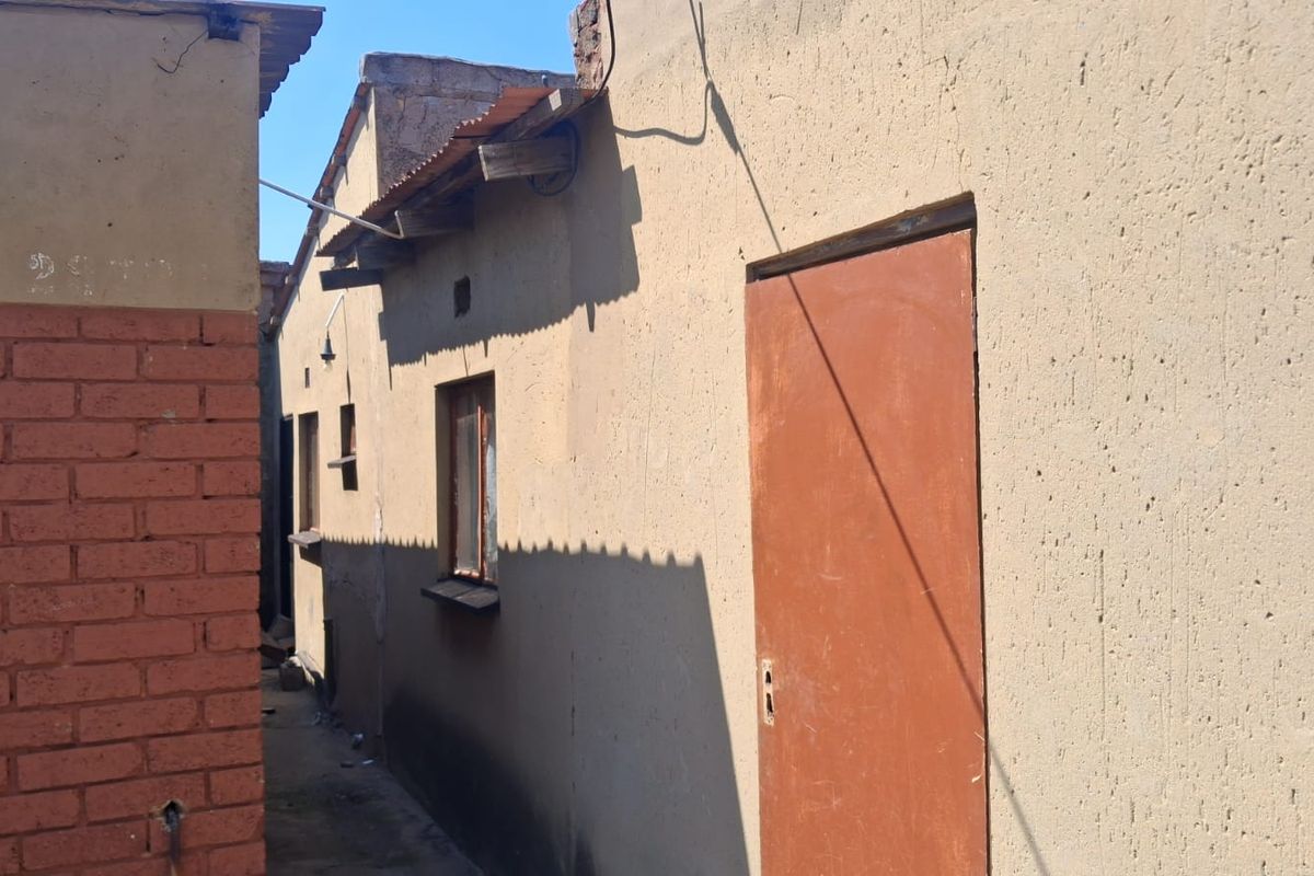A narrow exterior side passage showing the rear building elevation with plastered walls, steel door, and corrugated metal roofing.