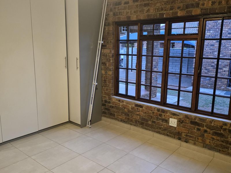 A bright bedroom featuring an exposed brick feature wall, tiled flooring, built-in cupboards, and a ceiling fan.