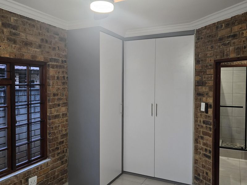 A clean, empty bedroom featuring exposed brick walls, built-in cupboards, tiled flooring, and a modern ceiling fan, with an adjacent bathroom entrance.