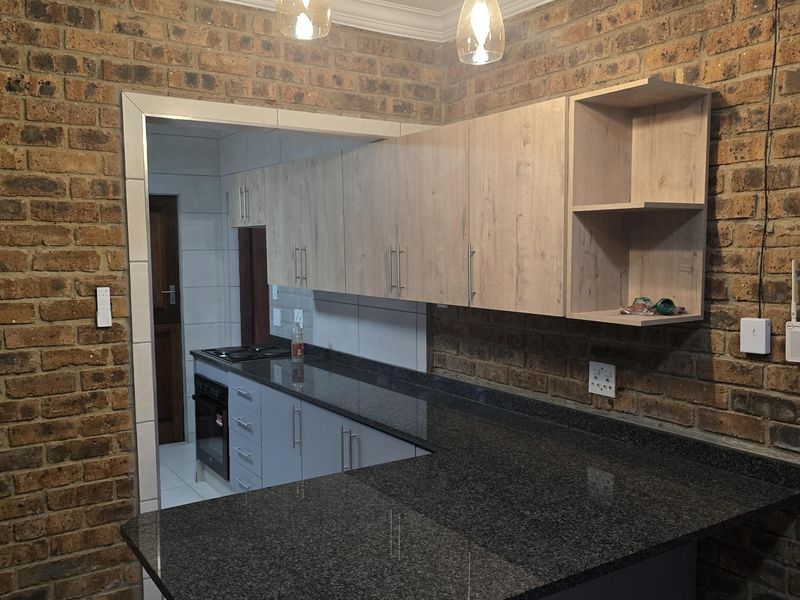Modern kitchen featuring exposed brick walls, granite countertops, light wood cabinetry, and a breakfast bar, with an adjacent scullery area visible.