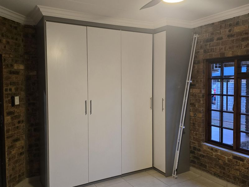 A bedroom featuring built-in cupboards, a ceiling fan, tiled flooring, and exposed brick walls, offering a clean and functional space.