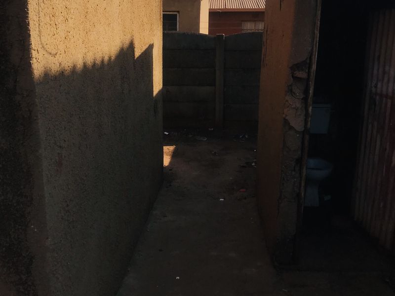 A narrow exterior passage between structures featuring a concrete pathway, boundary wall, and an external toilet facility with visible masonry deterioration.