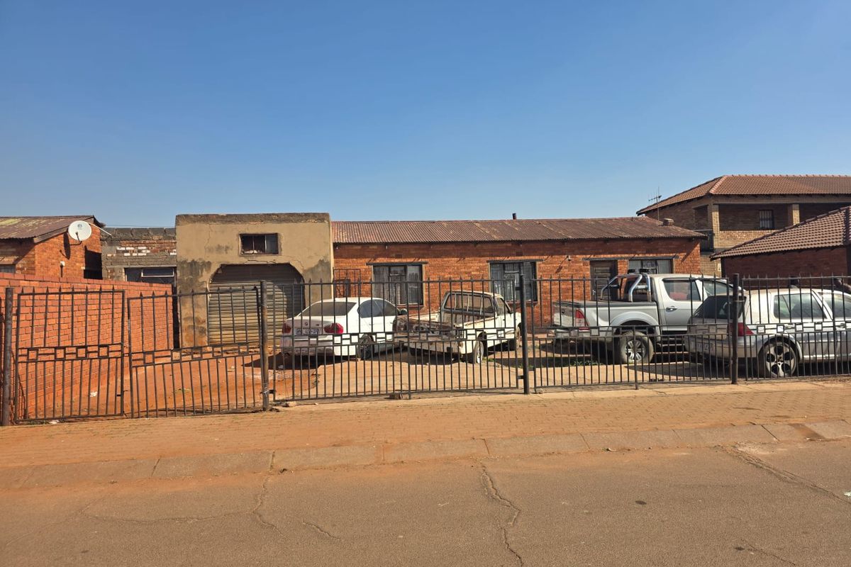 Front view of a single-story brick house featuring a metal security fence, garage access, and a corrugated metal roof.