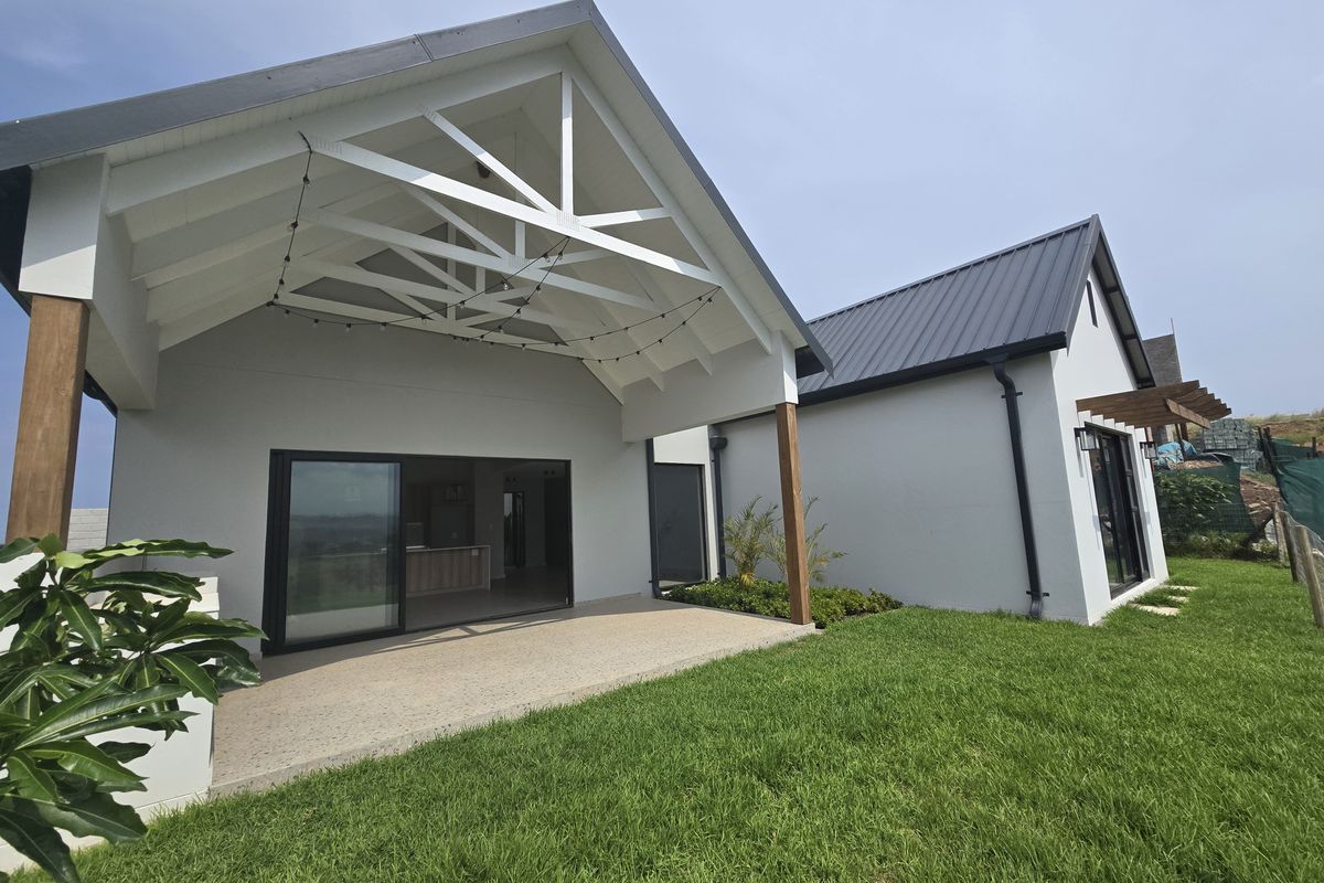 A modern rear exterior view featuring a spacious covered patio with exposed timber trusses, sliding glass doors, and a well-maintained lawn.