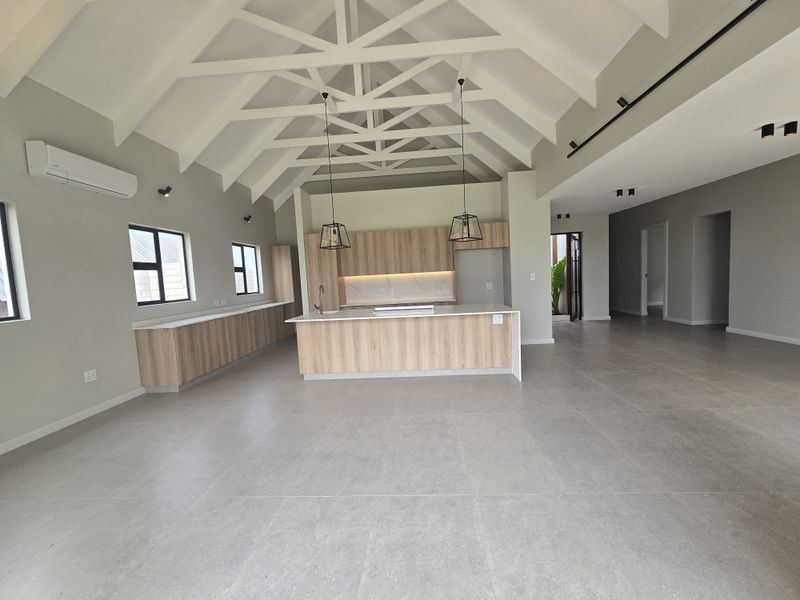 Spacious open-plan living and kitchen area featuring a high vaulted ceiling with exposed trusses, modern cabinetry, stone countertops, and large format floor tiles.