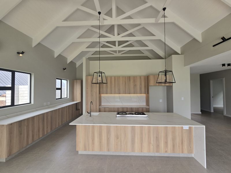 Modern kitchen featuring a high vaulted ceiling with exposed trusses, a central island with stone countertops, gas cooktop, and sleek wood-finish cabinetry.