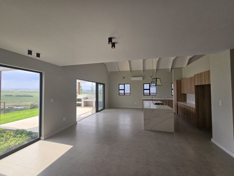 Spacious open-plan living and kitchen area featuring modern cabinetry, a stone-topped island, exposed rafters, and large sliding doors opening to a scenic outdoor patio.