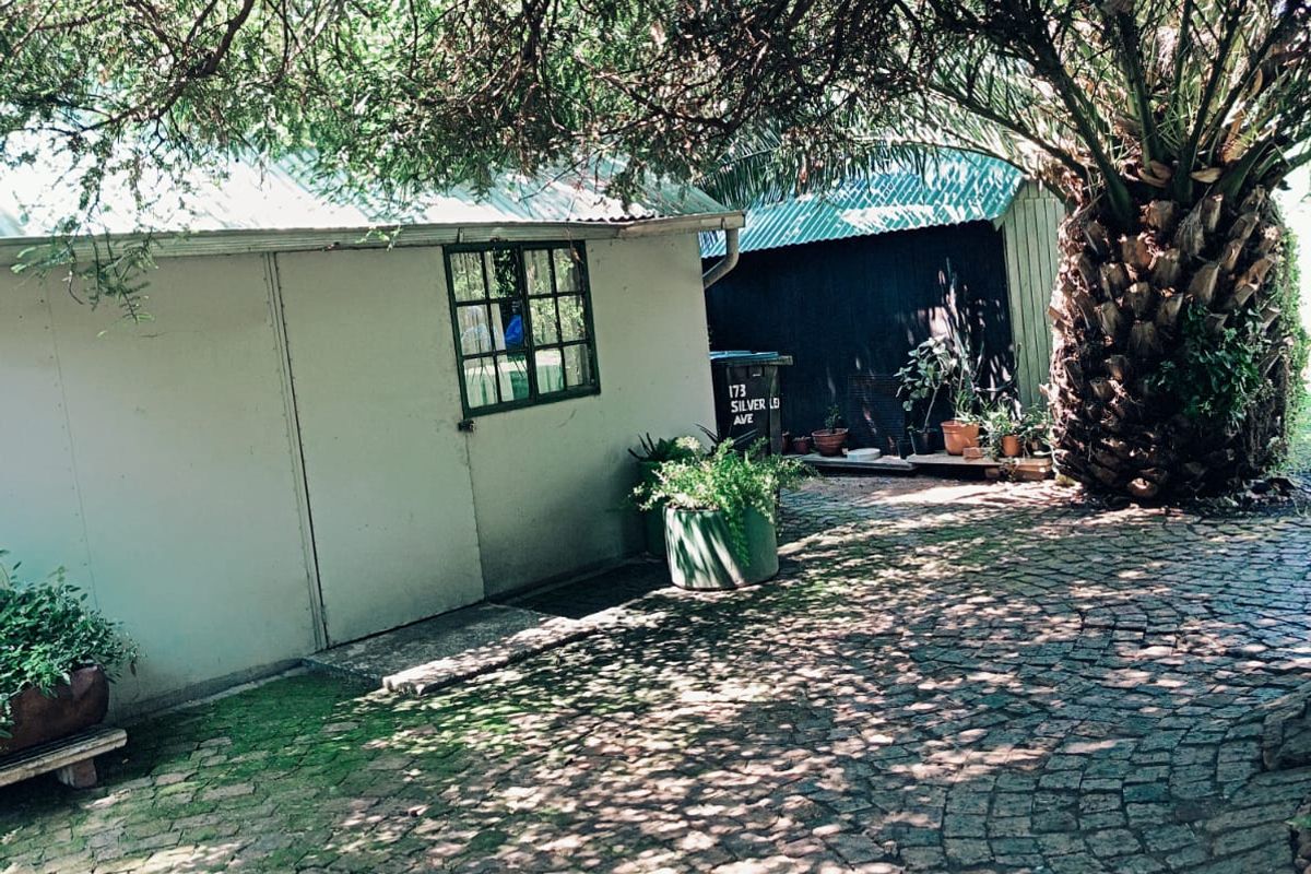 A rear exterior view featuring a cobblestone paved courtyard area with mature trees and a structure with a metal roof.