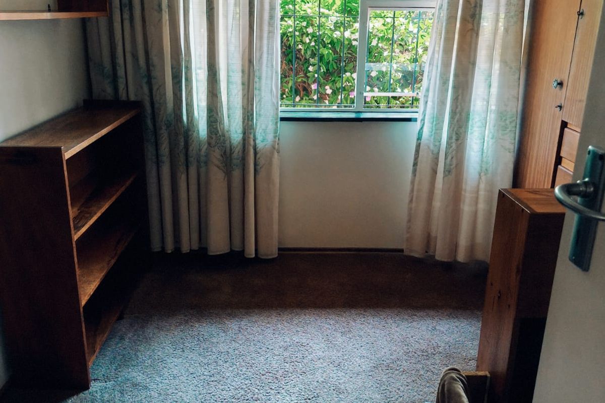 A bedroom featuring built-in wooden shelving and a wardrobe, with carpeted flooring and a window fitted with security bars.