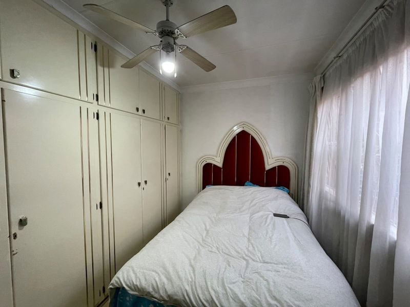 A compact bedroom featuring floor-to-ceiling built-in cupboards, a ceiling fan, and a window with curtains.