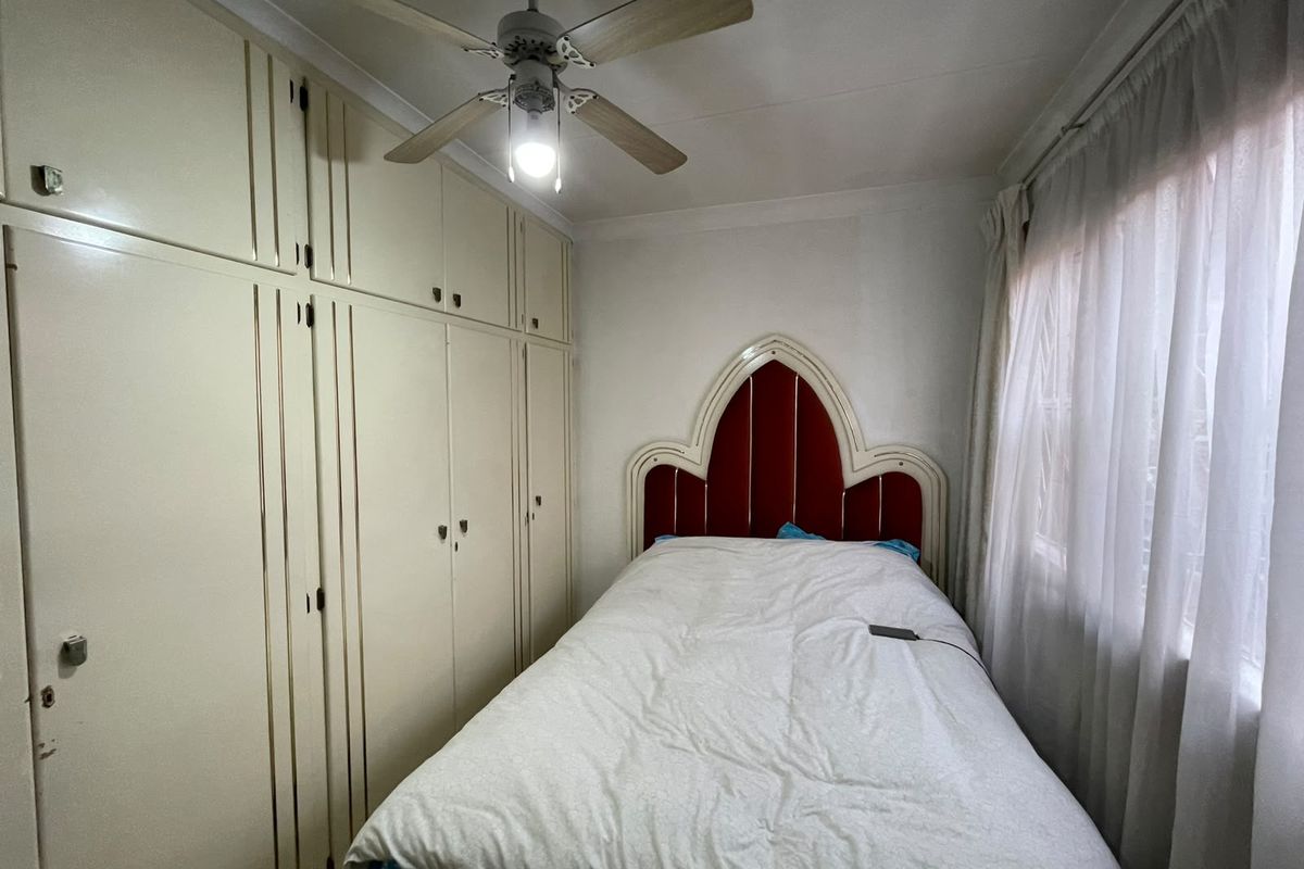 A compact bedroom featuring floor-to-ceiling built-in cupboards, a ceiling fan, and a window with curtains.