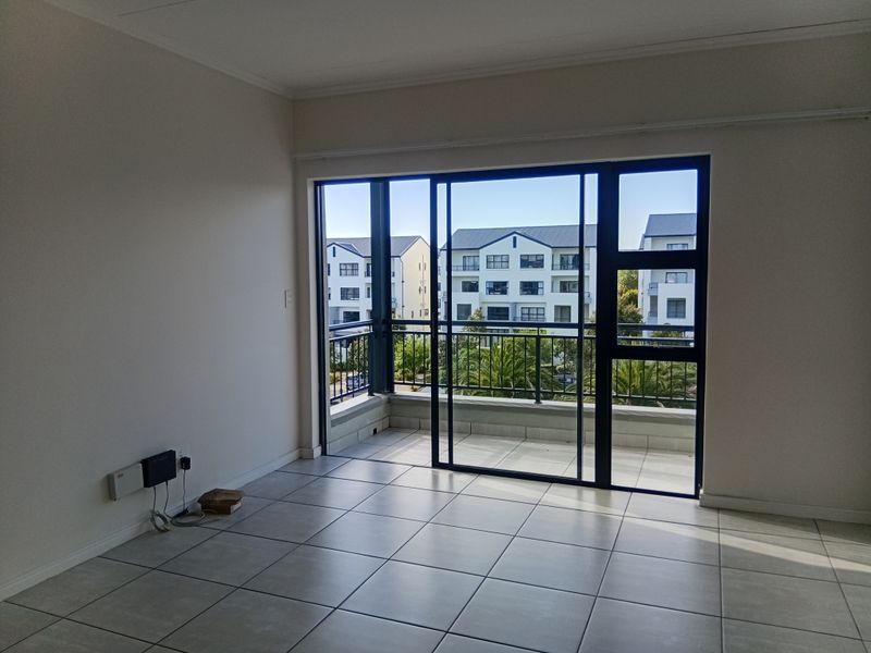 An empty living room featuring neutral tiled flooring and a large sliding glass door that opens onto a private balcony with neighbourhood views.