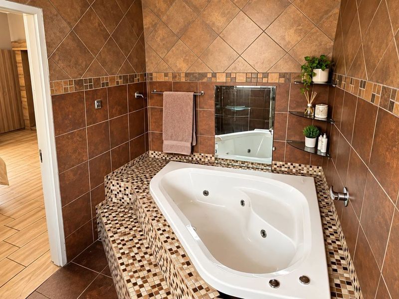 A bathroom featuring a corner spa bath with tiled surround and decorative mosaic accents, complemented by wall-mounted shelving and a mirror.