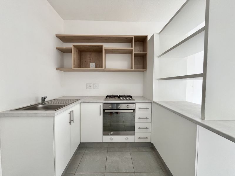 A clean, modern kitchen featuring white cabinetry, light wood open shelving, a built-in oven with gas hob, and durable tiled flooring.