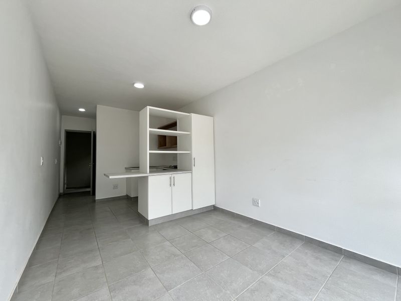 An empty open-plan living and kitchen area featuring neutral tiled flooring, built-in white cabinetry, and recessed ceiling lights.