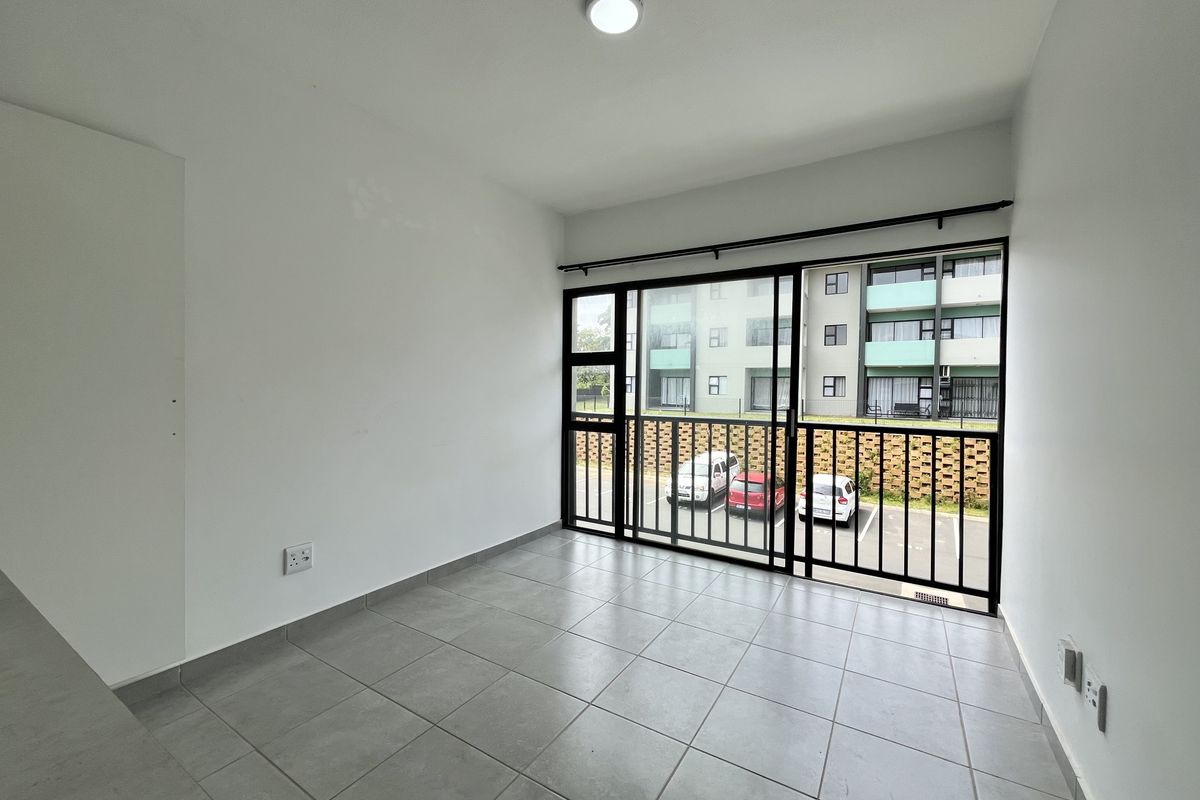 A bright, empty living area featuring neutral tiled flooring and a large sliding glass door with a security gate, offering views of the complex.