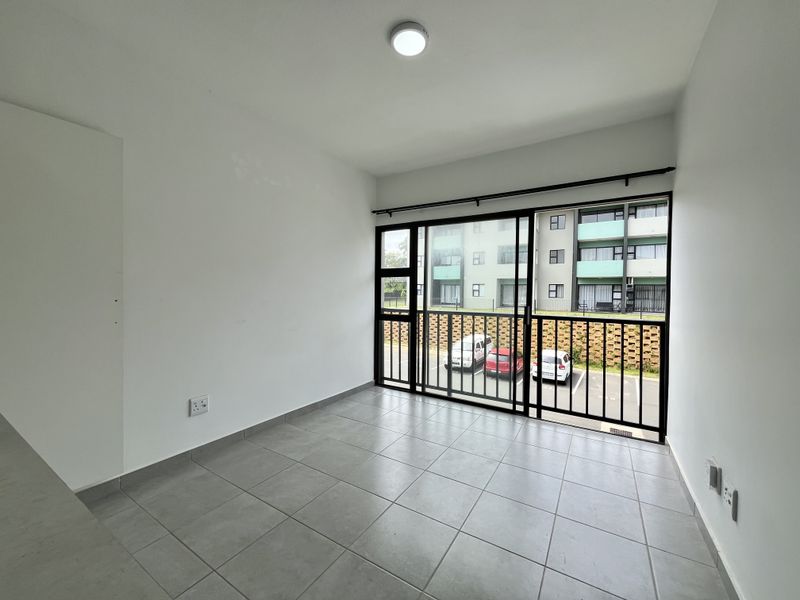 A bright, empty living area featuring neutral tiled flooring and a large sliding glass door with a security gate, offering views of the complex.