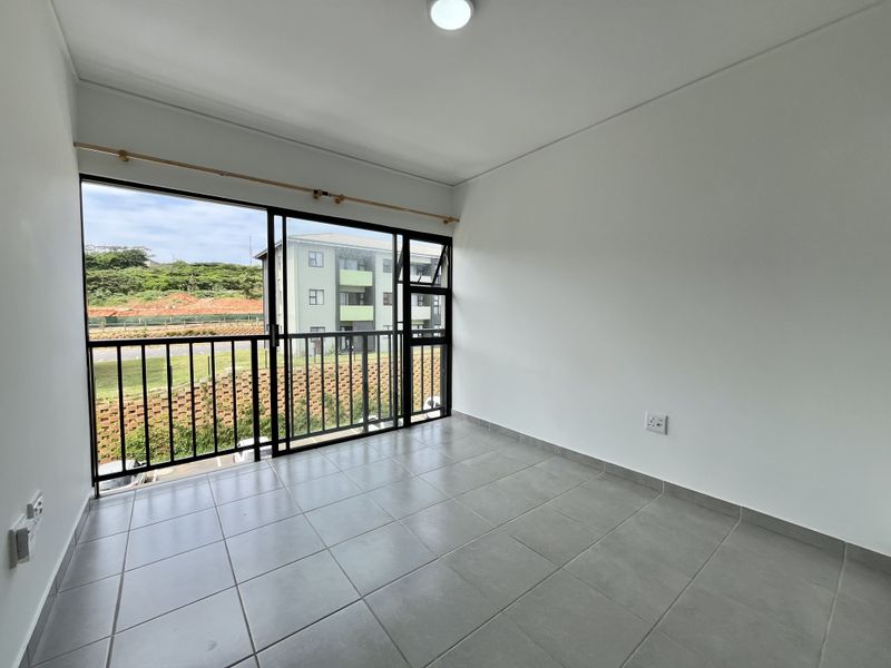 A bright, empty bedroom featuring neutral tiled flooring, a large sliding glass door leading to a balcony, and clean white walls.