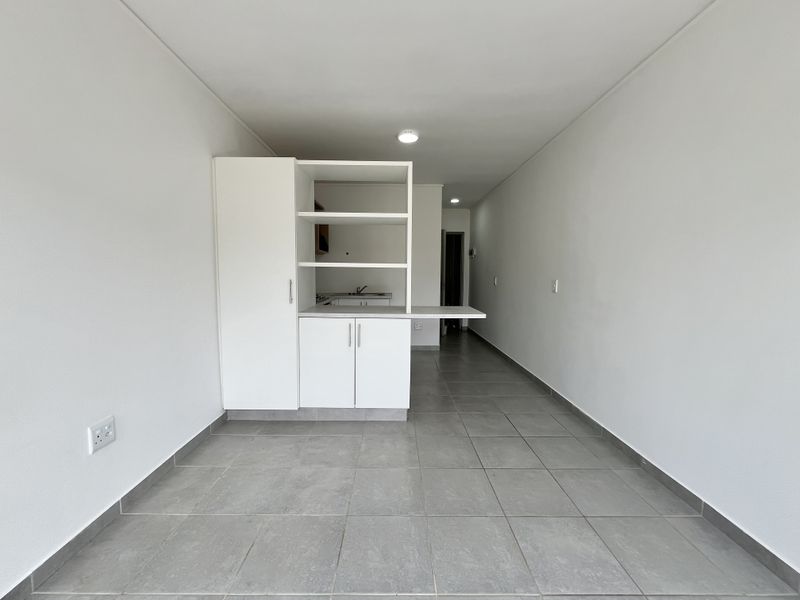 A bright, open-plan living and kitchen area featuring modern grey tiled flooring, clean white walls, and built-in cabinetry with integrated storage.