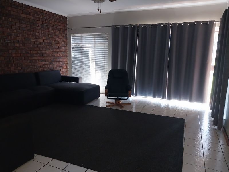A living room featuring a prominent exposed brick wall, tiled flooring, and large windows with blinds and curtains.