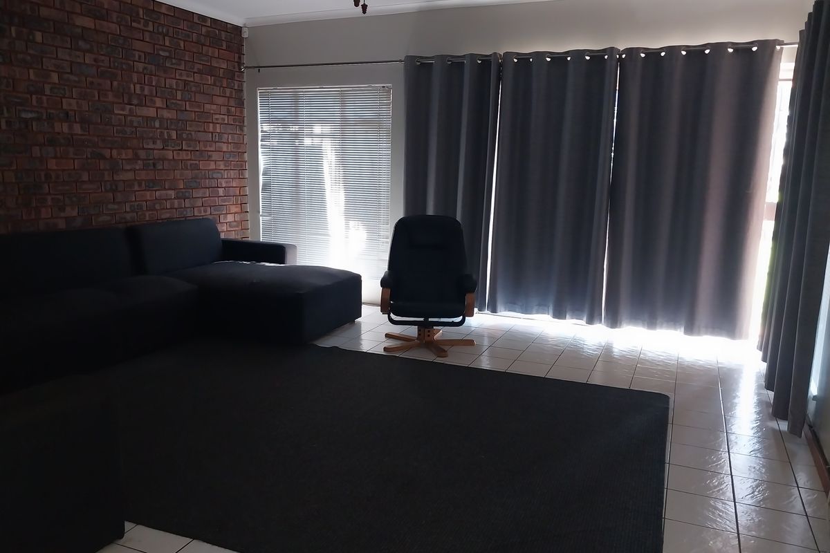 A living room featuring a prominent exposed brick wall, tiled flooring, and large windows with blinds and curtains.