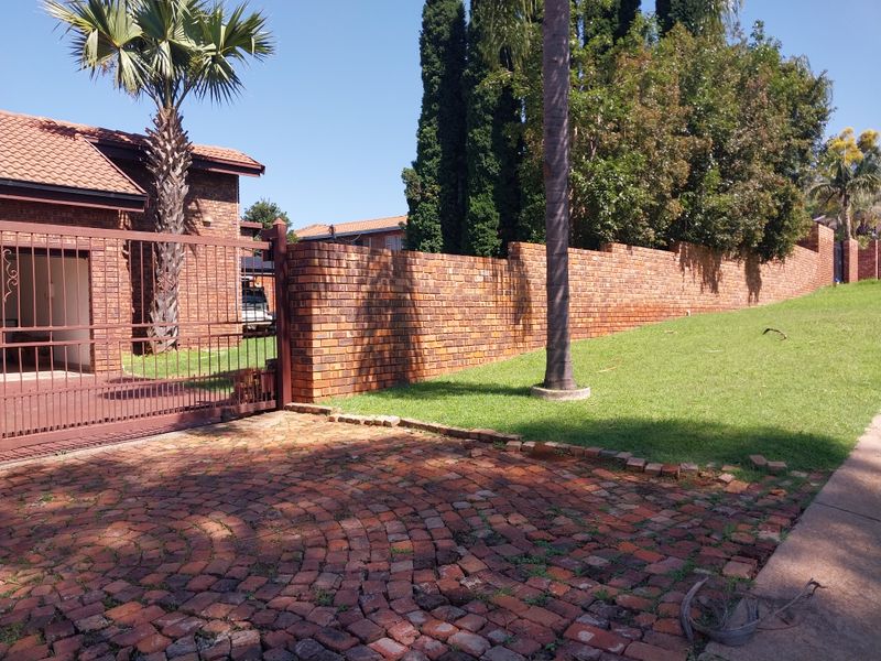 Front exterior view featuring a brick boundary wall, metal security gate, and a brick paved driveway leading to the property entrance.