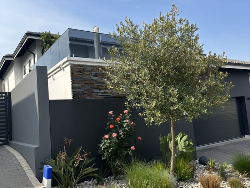 Modern cluster home exterior featuring a paved driveway, stone-clad accents, glass balustrade, and a well-maintained landscaped garden with an automated garage.