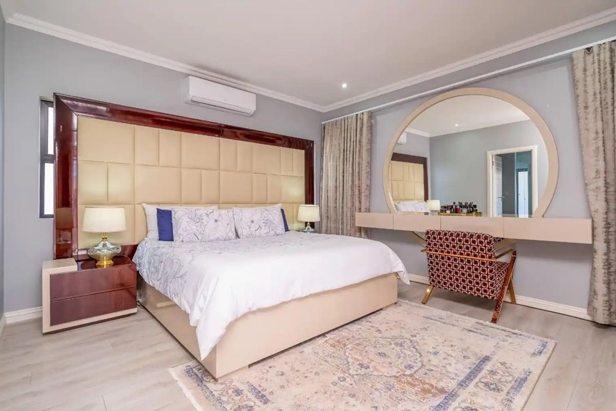 Spacious bedroom featuring a large built-in headboard, integrated vanity area with circular mirror, wall-mounted air conditioning, and modern laminate flooring.