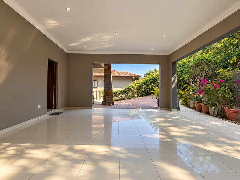 A spacious, covered patio featuring polished tile flooring, recessed lighting, and a decorative stone-clad pillar, offering a seamless transition to the garden area.
