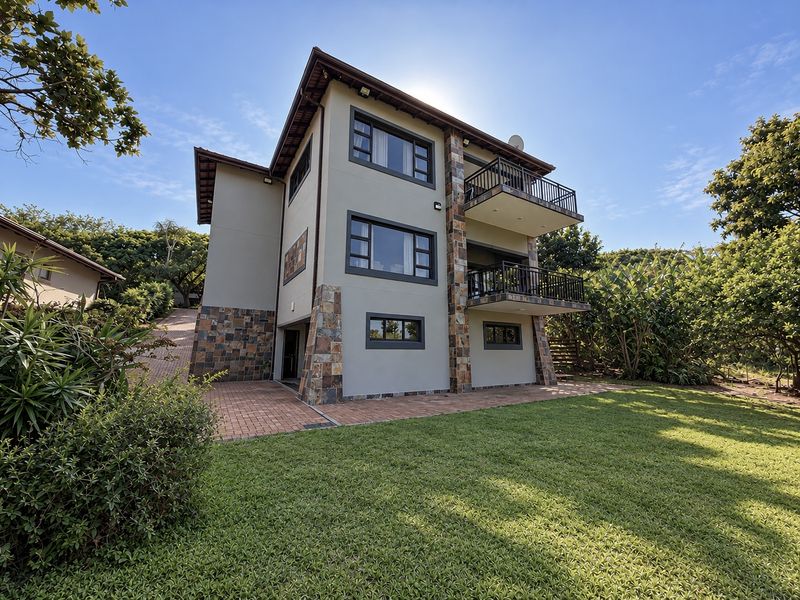 A well-maintained multi-story residential property featuring stone cladding, multiple balconies, a paved patio area, and a neatly landscaped garden.