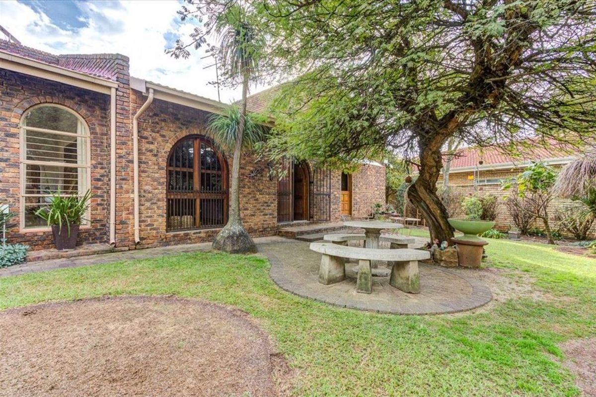 A rear exterior view of a face brick house featuring arched windows, doors, and a paved patio area set within a garden.