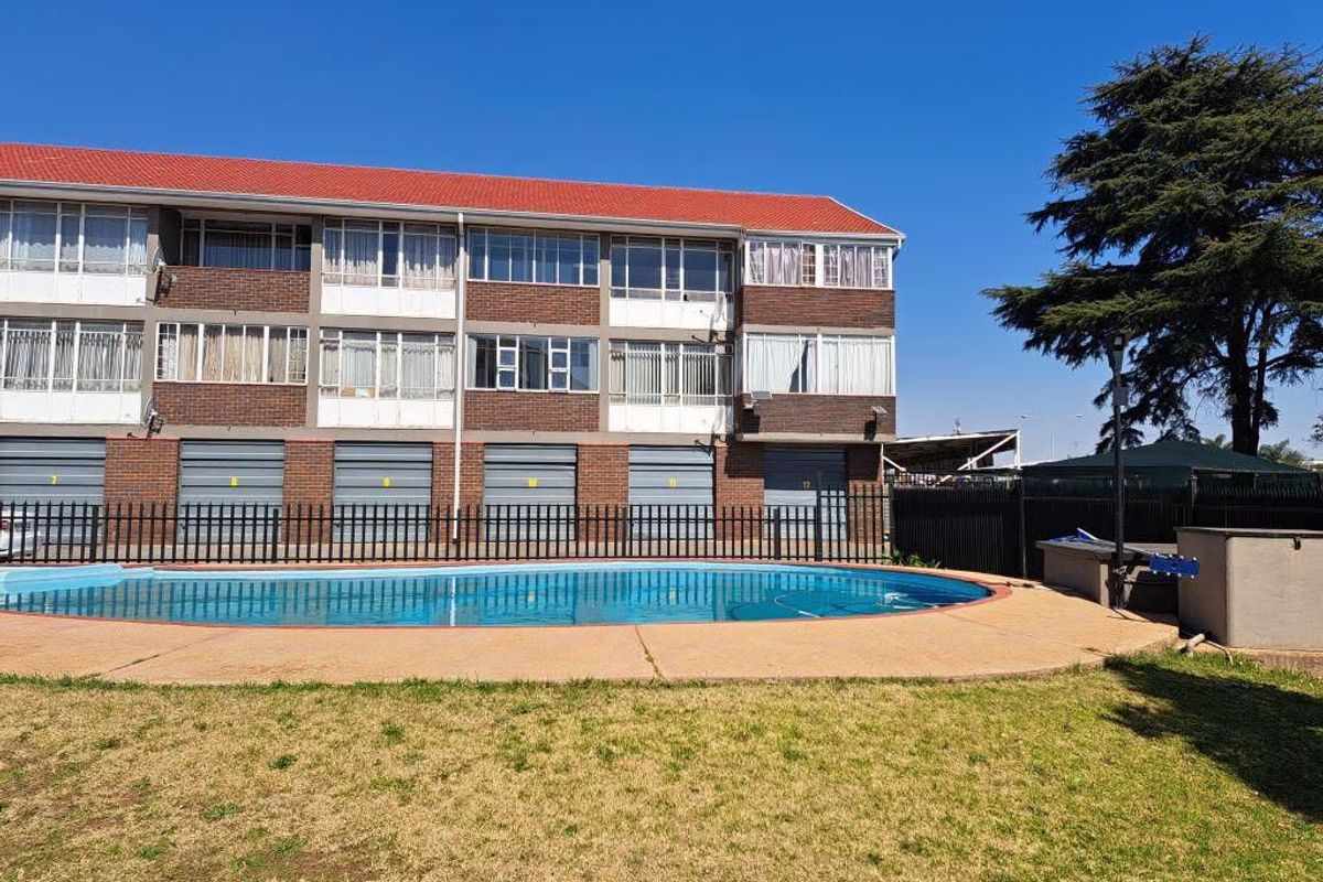 Exterior view of an apartment complex featuring a communal swimming pool, paved deck area, and ground-level garage units.